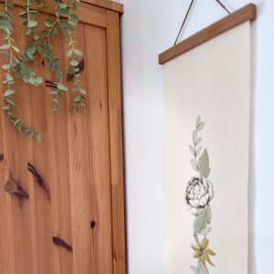 הדפסה ייחודית על בד קנבס בין מוטות עץ כפריים לתלייה על קיר באווירה כפרית וטבעית בפורמט צר ואורך מתאים לתליה על קיר צר במעברים עיצוב מיוחד עם המילים לראות את הטוב איור פרחים עדין לגובה מנטה חרדל מוקה חום