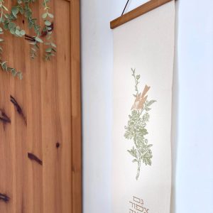 הדפסה ייחודית על בד קנבס בין מוטות עץ כפריים לתלייה על קיר באווירה כפרית וטבעית בפורמט צר ואורך מתאים לתליה על קיר צר במעברים עיצוב מיוחד עם המילים גם ציפור מצאה בית ודרור קן לה איור פרחים צמחים ירוק חום וציפור