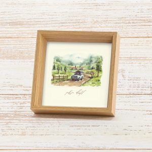 מסגרת תמונה ייחודית ריבוע עומד עם עובי להנחה על מדף עם פרינט של איור כמו צבעי מים עדין של נוף מאיר ושליו גווני ירוק שמנת תכלת בהיר עם רכב נוסע בשביל כפרי עם הכיתוב לאסוף רגעים